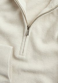 Vaaleanbeige pullover, jossa on neljännesvetoketju kauluksessa, teksturoitu kudottu kangas, ribbaatut hihansuut ja sileä vetoketju.