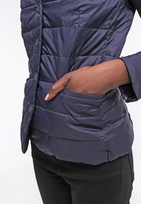 Veste matelassée bleu foncé avec des boutons sur le devant, montrant une main partiellement dans une poche latérale, portée avec un pantalon noir.