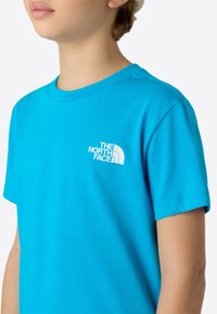 Turquoise cotton t-shirt with short sleeves, featuring a white embroidered logo on the left chest. Round neckline, smooth texture.