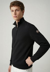 Black quarter-zip pullover made of soft fabric, featuring a high collar and ribbed cuffs. Includes a logo patch on the left sleeve.