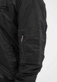 Black jacket with a textured fabric, featuring a zip pocket on the arm and a ribbed hem. Sewn logo visible on the chest.