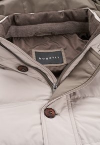 Beige Pufferjacke mit Reißverschluss, braunen Knöpfen, weichem Innenfutter und einem Etikett von Bugatti. Glatte, gesteppte Textur.