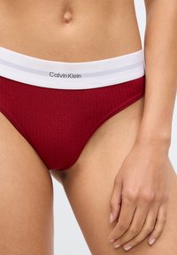 Punane ribadega bikiinipüksid, millel on valge elastne vöökoht koos "Calvin Klein" brändimisega. Suurendatud vaade kangale ja disainidetailidele.