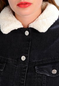 Jeansjacka i svart tyg, med en vit imitation av päls vid kragen och silverfärgade tryckknappar. Designen inkluderar en klassisk passform och fem fickor.