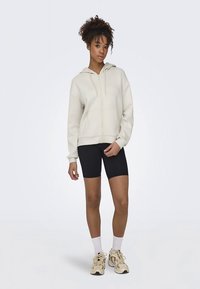 Lichtbeige hoodie met ritssluiting en een losse pasvorm, gecombineerd met zwarte fietsbroekjes. Model draagt witte enkelsokken en beige sneakers.