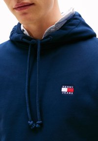 Marineblauwe hoodie van textuurstof met een trekkoordcapuchon, voorzien van een klein rechthoekig logopatch in rood, wit en blauw.