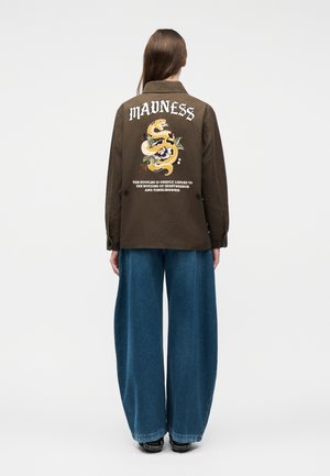 Personne portant une veste marron avec le texte "MADNESS" et un motif de serpent au dos, associée à un jean bleu à jambes larges et des chaussures noires.