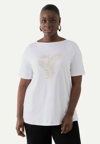 Camiseta de algodón blanca con un estampado de helecho dorado en el centro. La camiseta tiene mangas cortas y un escote redondeado, combinada con pantalones negros.