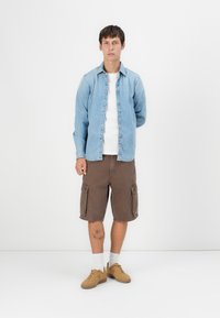 Lichtblauwe denim overhemd met knopen, wit T-shirt, bruin cargoshorts met zakken, tan sneakers en witte sokken. Neutrale achtergrond.