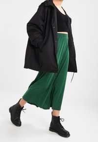 Chaqueta oversized negra, top corto negro, pantalones verdes de pierna ancha y botas negras con cordones. Las telas parecen suaves con un ajuste casual.