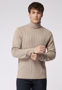 Beige Rollkragenpullover mit Strukturstrick und gerippten Bündchen sowie Saum. Weiche Textur, figurbetonte Passform und auffälliges Drehmuster.