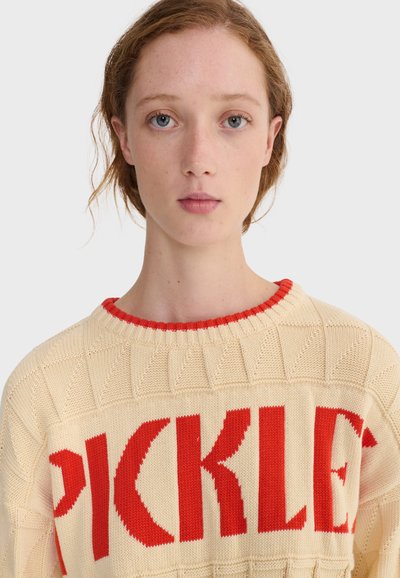 Jeune femme à la peau claire et aux yeux bleus portant un pull en tricot crème avec un grand texte rouge "PICKLES" sur la poitrine, fond neutre.