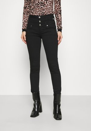 Kvinde iført sorte skinny jeans med høj talje og fire sølvknapper samt sorte ankelstøvler med hæl, kombineret med en leopardmønstret langærmet top.