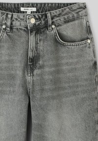 Un jean baggy en denim gris présente une texture usée avec des zones décolorées, un design à cinq poches, des accessoires en métal et des détails de couture subtils.