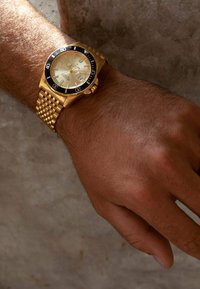 Goldene analoge Armbanduhr mit rundem Zifferblatt, beiger Anzeige und schwarzem Lünette. Das geflochtene Metallarmband hat eine strukturierte Oberfläche.