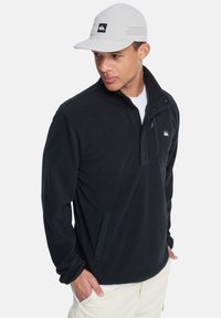 Sudadera de forro polar negra con medio botón en la parte delantera, bolsillo lateral y parche de logo. Combinada con una gorra gris claro con diseño perforado.
