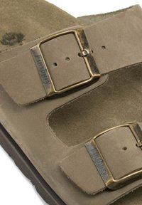 Sandale en daim marron dotée de deux boucles métalliques réglables, d'un design à bout ouvert et d'une semelle intérieure texturée. Semelle plate avec un léger détail en biais.