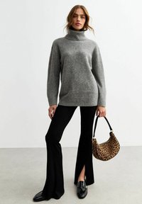 Grijze gebreide coltrui met lange mouwen, gecombineerd met zwarte flared broeken. Model houdt een tas met luipaardprint vast met een zwarte strap.