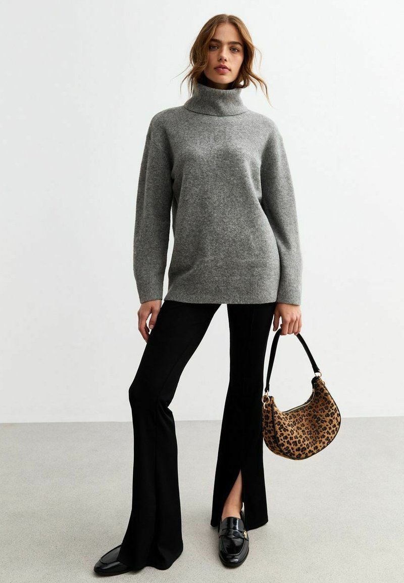 Grijze gebreide coltrui met lange mouwen, gecombineerd met zwarte flared broeken. Model houdt een tas met luipaardprint vast met een zwarte strap.