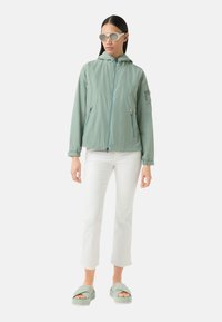 Lichtgroene, ritsjack met een capuchon, twee met rits afgesloten zakken, gestyled met witte cropped broek en groene slidesandalen.
