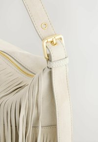 Blød beige suede taske med frynse-design, gyldne metaldele og lynlåslukning. Remmen har stikninger og metalring.
