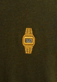 Una tela verde presenta un pequeño diseño bordado de un reloj amarillo, con una esfera rectangular y detalles texturizados en la correa.