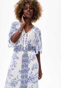 Witte jurk met blauwe bloemen- en geometrische patronen, v-hals, korte fladdermouwen en een strikdetail in de taille. Lichtgewicht materiaal.