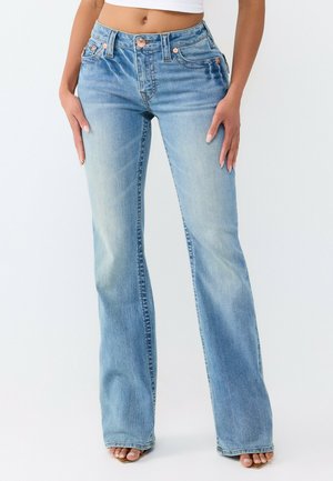 Femme portant un jean flare taille haute bleu clair et un top court blanc, debout, les mains sur les hanches, devant un fond uni.