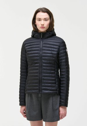 Donna con giacca nera trapuntata con cappuccio e pantaloncini grigi, in piedi davanti a uno sfondo bianco semplice.