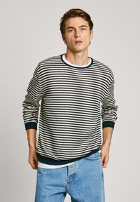 Suéter a rayas en crema y marino con cuello redondo, puños y dobladillo acanalados. Lleva encima una camiseta blanca; combinado con unos vaqueros azul claro.