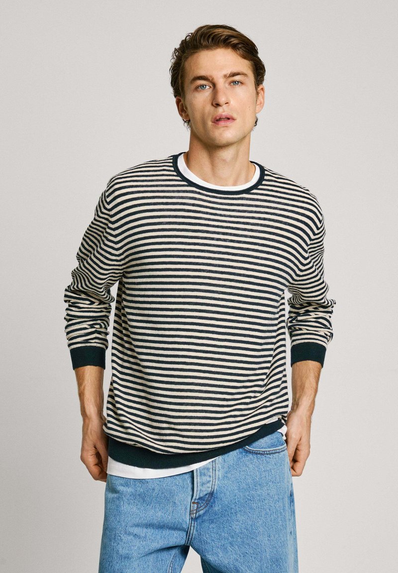 Suéter a rayas en crema y marino con cuello redondo, puños y dobladillo acanalados. Lleva encima una camiseta blanca; combinado con unos vaqueros azul claro.