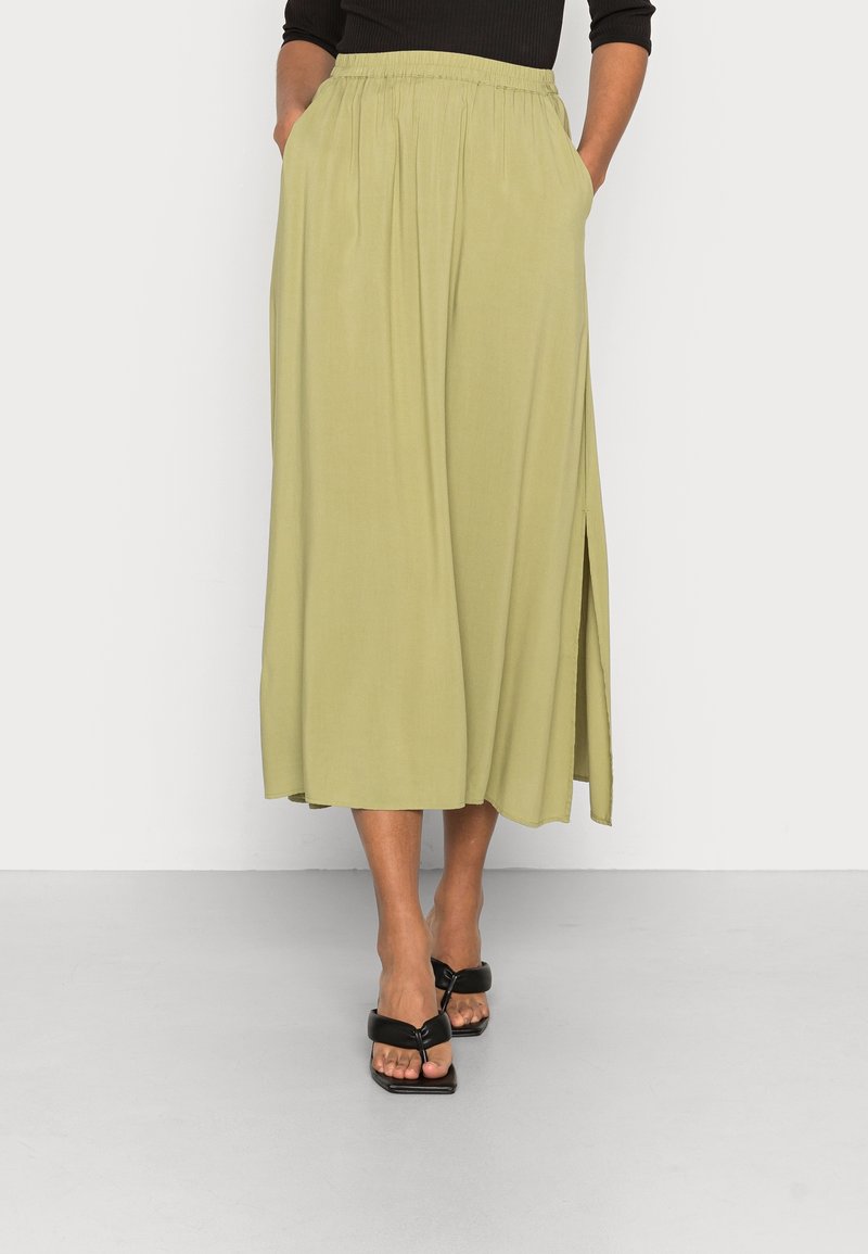 Femme portant une jupe midi vert clair avec une fente latérale, un haut noir et des sandales noires à talons ouverts, debout les mains dans les poches.