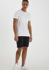 T-shirt en coton blanc avec un léger motif de palmier, associé à un short noir et des baskets blanches pour un look décontracté.