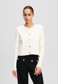 Cardigan corto di colore crema realizzato in tessuto morbido, con maniche lunghe e bottoni rotondi color oro lungo il davanti, abbinato a pantaloni neri.