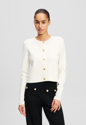 Cropped cream cardigan made of soft fabric with long sleeves and gold-tone round buttons down the front, paired with black pants.