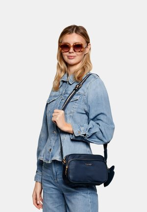 Donna con giacca di jeans e jeans che indossa occhiali da sole con montatura tartarugata, tiene una borsa a tracolla blu navy con cerniera dorata e logo sulla parte anteriore.