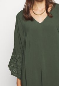 Olivgrünes Tunika mit V-Ausschnitt und ausgestellten Ärmeln, versehen mit filigranen Spitzenverzierungen an den Bündchen, aus einem glatten, leichten Stoff gefertigt.