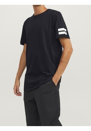 Mand iført en ensfarvet sort T-shirt med korte ærmer med to hvide vandrette striber på venstre ærme og sorte bukser, hånden i lommen.