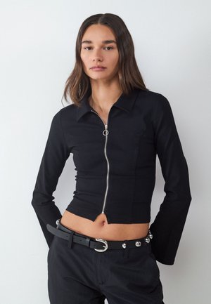 Donna che indossa un top nero aderente a maniche lunghe con cerniera e pantaloni neri con cintura nera con borchie argento e fibbia argentata.