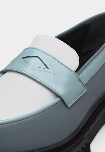 Δίχρωμο δερμάτινο μοκασίνι με λευκό επάνω μέρος, ανοιχτό μπλε λουράκι με διαμάντι σχήματος κοπή και μαύρη σόλα με ριγέ άκρη.