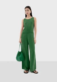 Une combinaison verte avec un corsage ajusté et un pantalon large, fabriquée en matériau léger. Associée à un sac à cordon vert et des sandales plates.