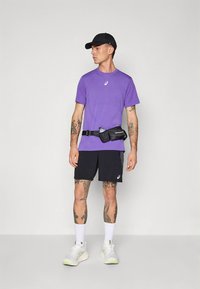 Lilla lühikeste varrukatega spordisärk logoga, mustad lühikesed püksid ja must vöökoha kott. Valged tossud ja must nokamüts täiendavad rõivastust.