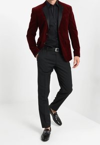 Röd sammetskavaj med svarta slag, kombinerad med svart skjorta och skräddarsydda svarta byxor. Svarta lackskor fulländar outfiten.