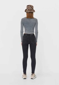 Grå långärmad topp i kombination med svarta tajta jeans. Anmärkningsvärd leopardmönstrad bucket-hatt. Modell sedd bakifrån, iförd beigefärgade sneakers.