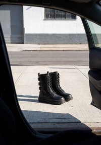 Een paar zwarte veterkistens op een stoep, gezien door een open autodeur met een straat en gebouw op de achtergrond.