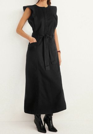 Femme portant une robe midi noire sans manches avec épaules matelassées, ceinture nouée à la taille, poches avant et bottines noires à bout pointu et talons.