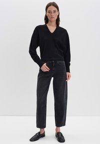 Frau in schwarzem V-Ausschnitt-Pullover, hoch taillierten schwarzen Jeans und schwarzen Slippern, die vor einem schlichten weißen Hintergrund steht.