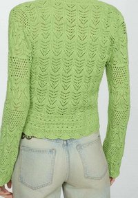Pull vert en maille à manches longues, avec un motif ajouré et un bord inférieur en festons. Porté avec un jean bleu clair.