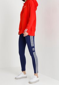 Sudadera con capucha roja con bolsillo canguro y logo blanco, combinada con mallas azul marino con tres franjas blancas y logo de Adidas, completada con zapatillas blancas.
