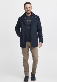 Mornarski kaput od vune s mješavinom, dvostrukim kopčanjem, detaljima na dugmadi i bočnim džepovima, uparen s tamnim duksericom i khaki hlačama.